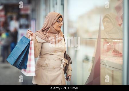 Donna musulmana di mezza età che indossa hijab con un volto felice sta camminando in ambiente urbano, andando allo shopping che tiene borse colorate di carta. Foto Stock