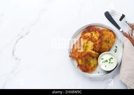 Frittelle di patate. Frittelle di patate fritte fatte in casa o latkes con crema e cipolle verdi in piatto rustico su sfondo bianco tavolo di marmo. Stile rustico. Foto Stock