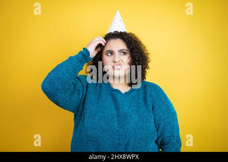 Giovane bella donna che indossa un cappello di compleanno sopra isolato sfondo giallo confondere e meraviglia circa la domanda. Incerto con il dubbio, pensando con ha Foto Stock