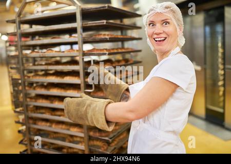 Felice giovane donna panettiere apprendista spingendo carrello ripiano con prodotti freschi cotti in panetteria Foto Stock