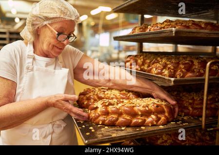 Donna anziana come panettiere con una pagnotta di lievito appena sfornata con mandorle nella panetteria Foto Stock