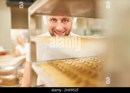Sorridente giovane uomo panettiere in scaffale con prodotti da forno in panetteria Foto Stock