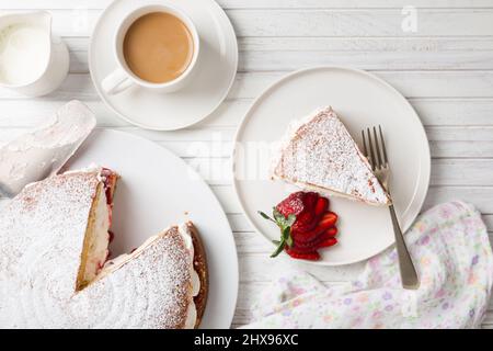 Vista dall'alto di un pan di Spagna Vittoria fatto in casa riempito con fragole, marmellata e panna montata su un tavolo di legno bianco e una tazza di caffè o tè Foto Stock
