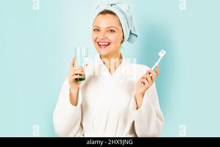 Donna in accappatoio e asciugamano sulla testa spazzolando i denti trattiene l'acqua di vetro. Routine mattutina. Cura dentale. Foto Stock