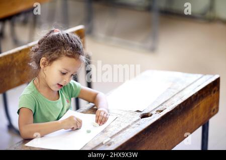 Esprimendo la sua creatività. Ragazza piccola carina seduta ad una scrivania della scuola e disegno. Foto Stock