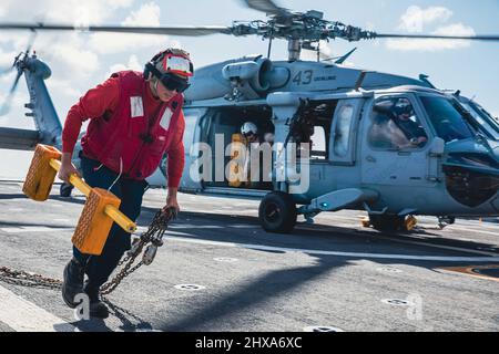 220308-N-GF955-1151 MAR DEI CARAIBI - (8 MARZO 2022) -- Mineman Seaman Apprentice Aizik Ramirez, assegnato alla nave da combattimento litoraneo Freedom-Variant USS Billings (LCS 15), rimuove i cunei e le catene da un elicottero MH-60s Sea Hawk assegnato alla “Shadow Det” di Helicopter Sea Combat Squadron (HSC) 28, distaccamento 7, durante le operazioni di volo, 8 marzo 2022. Billings è dispiegato nell'area delle operazioni della flotta USA 4th per sostenere la missione della Joint Interagency Task Force South, che include missioni di lotta contro il traffico illecito di droga nei Caraibi e nell'Est del Pacifico. (STATI UNITI Navy foto di massa Foto Stock