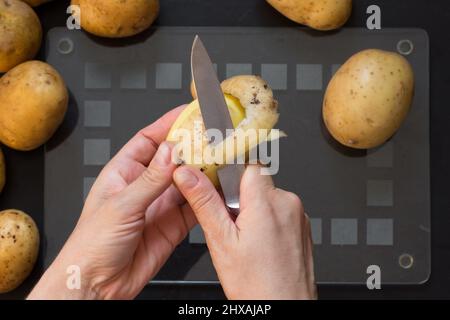 vista dall'alto della donna che pelava la patata cruda con il coltello sullo sfondo nero Foto Stock