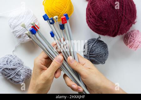 Le mani della donna tengono molti aghi di maglia di numeri diversi per scegliere lo spessore dei fili su sfondo bianco Foto Stock