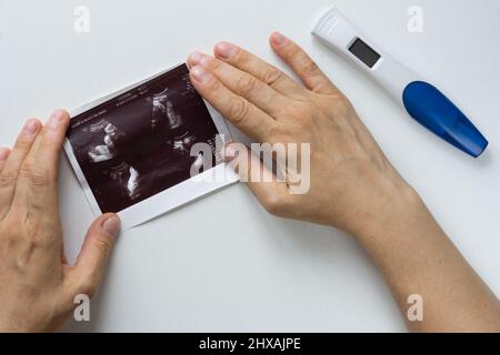 Le mani della donna tengono l'immagine dell'ecografia dell'embrione sullo sfondo bianco e sul test gravido Foto Stock