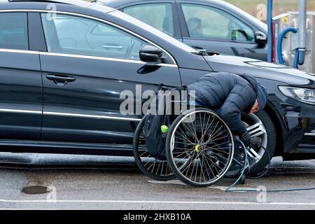 Riga, Lettonia, 4 gennaio 2021: Il conducente handicappato su sedia a rotelle pompa gli pneumatici della sua auto dal compressore in una stazione di benzina Foto Stock