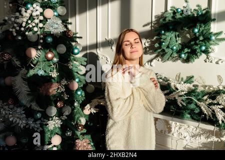 Una bella giovane donna con capelli biondi si erge sullo sfondo di un albero di Natale. L'anno nuovo è in arrivo. Atmosfera natalizia in un ambiente accogliente Foto Stock