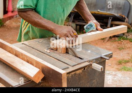 Un costruttore di cabinate con un pennel tra le sue dita sostiene un pezzo di legno come lui usa una vecchia sega elettrica per tagliare la sua estremità Foto Stock