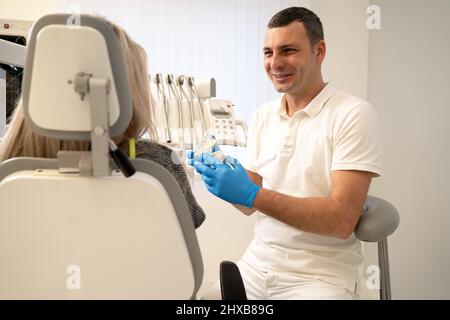 un uomo in un dentista medico di smok mostra un morso artificiale della mandibola di plastica per una paziente seduta nell'ufficio del dentista nell'ufficio vicino ai denti Foto Stock