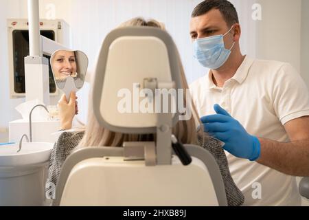 medico dentista in guanti blu che reggono un tavolo di tonalità denti, scegliendo il giusto colore smalto per la paziente femminile durante il trattamento dentale nella c Foto Stock