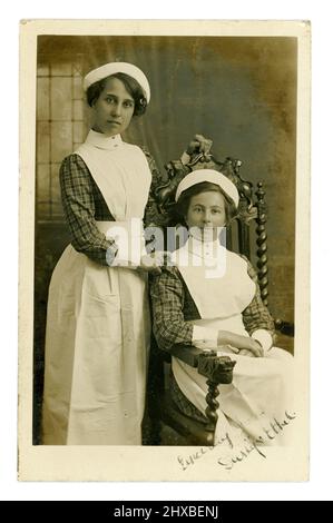 Ritratto originale da cartolina dell'era WW1 di 2 / due giovani infermiere, o infermiere in grembiuli amidati con bavaglino, berretti e polsini e collari staccabili. Entrambi indossano camicette controllate corrispondenti, così sono il personale che lavora nella stessa casa di cura, ospedale o grande casa privata. Le ragazze hanno firmato Daisy ed Ethel sul fronte, nell'area di Londra, nel Regno Unito intorno al 1915. Foto Stock