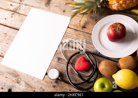 Vista ad angolo alto di vari frutti con stetoscopio e forma del cuore da carta bianca su tavola di legno Foto Stock
