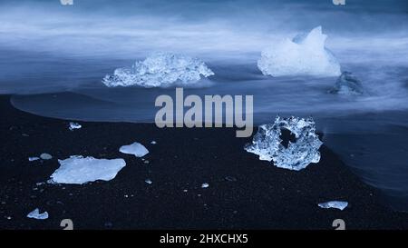 Ghiaccio sulla spiaggia nera vicino alla laguna del ghiacciaio Jökulsárlón, onde a lunga esposizione, atmosfera serale, ora blu, Parco Nazionale Vatnajökull, Islanda Foto Stock