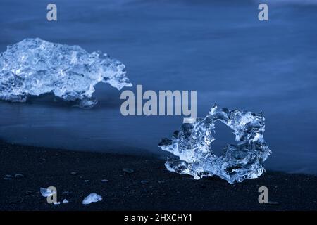 Ghiaccio sulla spiaggia nera vicino alla laguna del ghiacciaio Jökulsárlón, onde a lunga esposizione, atmosfera serale, ora blu, Parco Nazionale Vatnajökull, Islanda Foto Stock