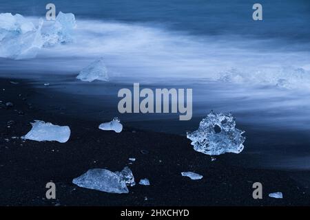 Ghiaccio sulla spiaggia nera vicino alla laguna del ghiacciaio Jökulsárlón, onde a lunga esposizione, atmosfera serale, ora blu, Parco Nazionale Vatnajökull, Islanda Foto Stock