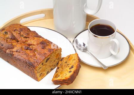 Tazza bianca e piattino con bordo decorativo in argento contenente caffè nero, su vassoio di legno con torta di frutta in primo piano. Foto Stock