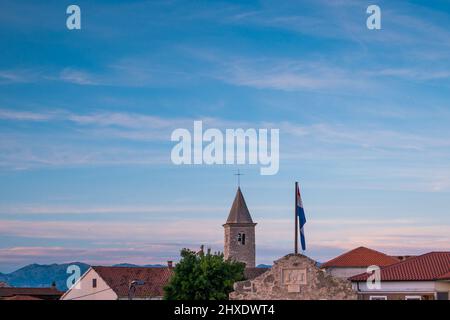 Skyline della città di Nin in Croazia con torre della chiesa, tetti rossi e bandiera croata su un cielo blu con piccole nuvole Foto Stock