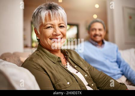 Abbiamo fatto bene. Ritratto di una donna più anziana felice che si rilassa sul divano con il marito sullo sfondo. Foto Stock