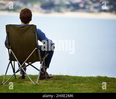 Il luogo perfetto per godersi il paesaggio. Vista posteriore di un giovane seduto su una sedia da campeggio e guardando la vista. Foto Stock