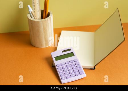 Notebook, calcolatrice, portamatite su una scrivania arancione. Sfondo giallo a parete. Spazio di lavoro Foto Stock