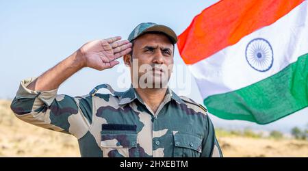 Vista frontale basso angolo di orgoglioso militare indiano saluta mentre sventola bandiera indiana sullo sfondo - concetto di patriota, nazionalismo Foto Stock