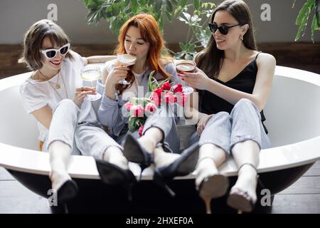 Tre giovani amiche adulte che si siedono insieme con bevande alcoliche nella vasca da bagno, hanno una festa di gallina o festeggiano le vacanze delle donne a casa Foto Stock