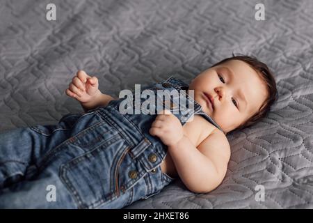 Ritratto del bambino grazioso che indossa il romper del denim che riposa sulla coperta grigia in casa. Il bambino assonnato si adagia sul letto in camera da letto ed esplora il mondo. Grigio Foto Stock