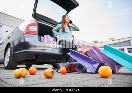 Vista ravvicinata e dal basso della frutta d'arancia sul terreno, sparsa dal pacchetto shopping. Sul fondo sfocato auto con bagagliaio aperto, dentro di essa seduta donna incinta che parla al telefono. Foto Stock