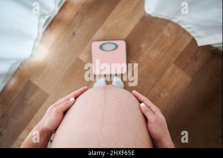 Foto ritagliata del ventre grande di donna incinta in piedi in casa davanti alle bilance da pavimento per pesare. Controllo della pesatura del peso corporeo durante la gravidanza prima del parto. Foto Stock