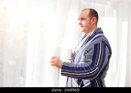 Un uomo calvo vestito con un housecoat bianco, grigio, nero e blu a strisce sta tenendo una tazza di caffè mentre tira da parte una tenda per sbirciare fuori la finestra. S Foto Stock