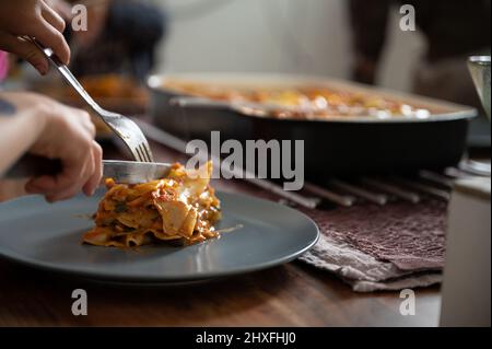 Primo piano vista di un bambino a mano tagliando deliziose lasagne di verdure vegane fatte in casa con un vassoio intero di esso sullo sfondo. Foto Stock