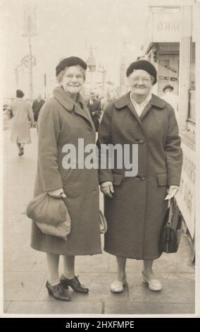 Due donne anziane in posa per un cameraman di strada, mare, 1950s. Sorridono alla macchina fotografica, vestiti con spesso cappotti che indossano cappelli. Venduto a loro su una cartolina da portare a casa. Foto Stock