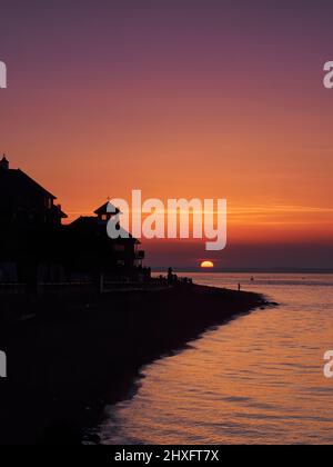 Un tramonto riccamente colorato visto da Cowes Beach, con il disco solare già in parte sotto l'orizzonte e la gente di sagome sulla spiaggia. Foto Stock