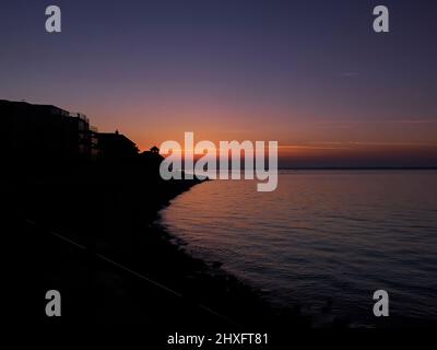 Un tramonto riccamente colorato visto da Cowes Beach, la luce del sole che passa attraverso le finestre di un edificio sul mare e le persone in silhouette sulla spiaggia. Foto Stock