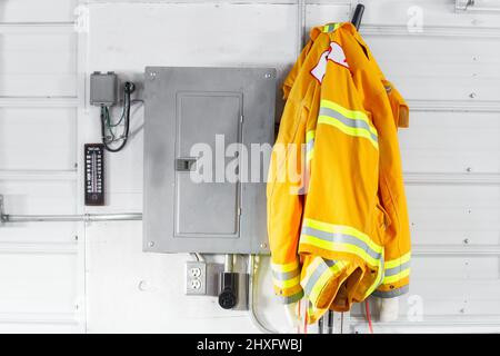 Un cappotto giallo per vigili del fuoco per adulti appeso accanto a una scatola elettrica su una parete di stagno Foto Stock