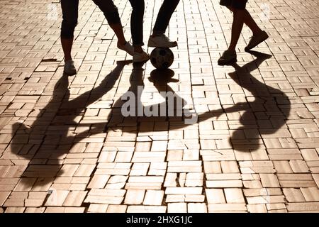 Calciarlo con alcuni amici. Immagine ritagliata delle gambe che calcia una palla con ombre sulla strada asfaltata. Foto Stock