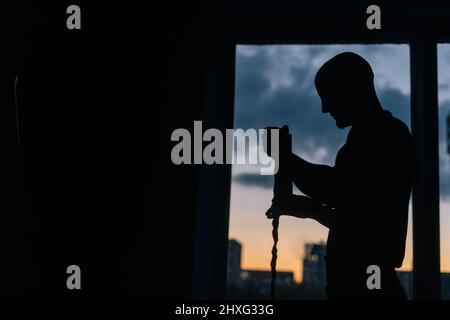 Silhouette di irriconoscibile boxer muscolare che avvolge il nastro da boxe intorno al polso in piedi sullo sfondo della finestra la sera al tramonto. Foto Stock