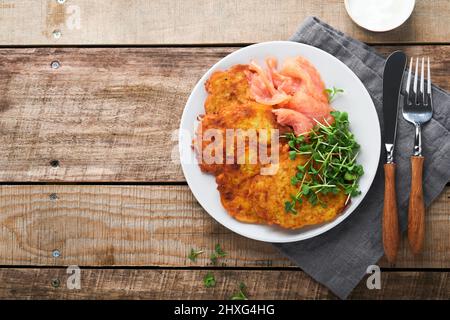 Frittelle di patate. Frittelle di patate fatte in casa fritte o latkes con crema, cipolle verdi, microgreens, salmone rosso e salsa in piatto rustico su vecchio legno ta Foto Stock