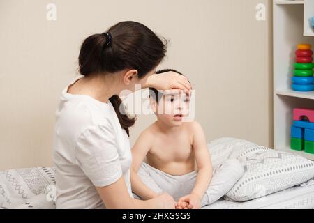 La madre misura la temperatura di un bambino malato, mette la mano sulla fronte. Malattie con febbre nei bambini, influenza o coronavirus Foto Stock
