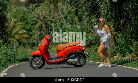 Donna su scooter rosso in abiti bianchi guida su strada forestale. Viaggio di ballo. Una ragazza caucasica turista in occhiali da sole danza, relax, riposo vicino a motore Foto Stock