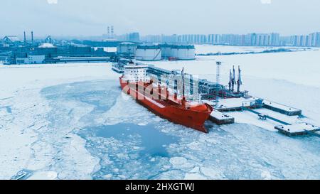 Terminal petrolifero settentrionale nella neve e in petroliera illuminata da luci rosse ormeggiate sull'acqua dell'oceano coperta di ghiaccio in vista aerea al crepuscolo della sera Foto Stock