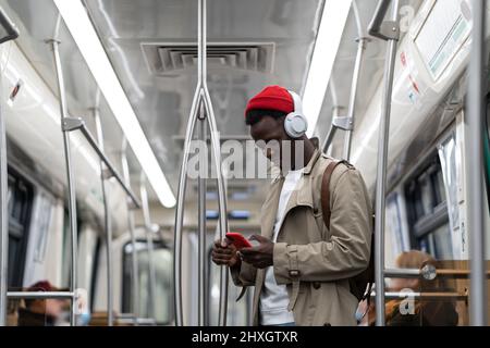 L'uomo hipster nero indossa cuffie wireless, ascolta la musica, utilizzando lo smartphone in treno della metropolitana Foto Stock