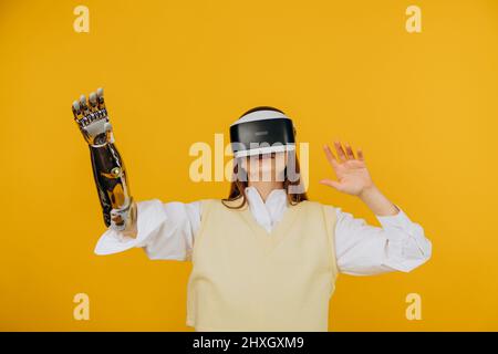 Donna concentrata in gilet con moderno braccio bionico e visore per realtà virtuale gioca il gioco su sfondo giallo primo piano Foto Stock