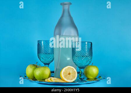 Ancora vita di una bottiglia di limonata e bicchieri circondati da mele e limoni su un vassoio di metallo su sfondo blu Foto Stock