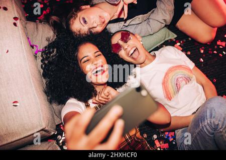 Selfie vibranti per persone vibranti. Vista dall'alto di tre amici felici che prendono un selfie mentre si trova sul pavimento ad una festa della casa. Gruppo di allegro Foto Stock