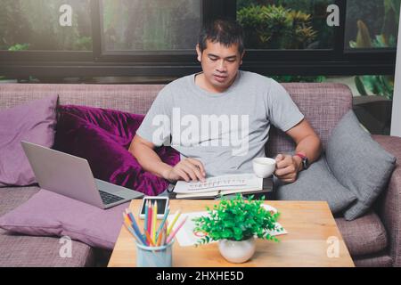 Uomo asiatico che tiene una tazza di caffè e lavora mentre si siede sul divano in ufficio domestico. Foto Stock